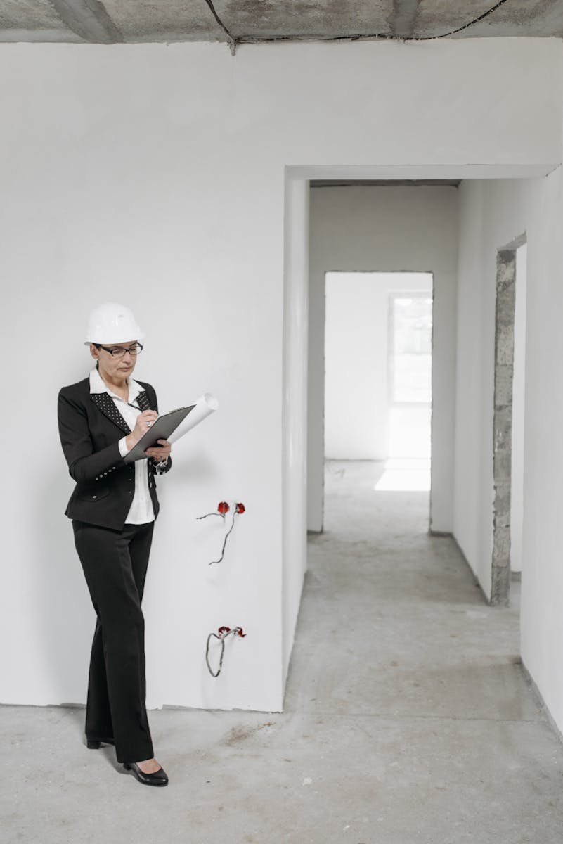 Photo by Pavel Danilyuk Female architect in hard hat inspecting an unfinished building interior with formal attire and clipboard.